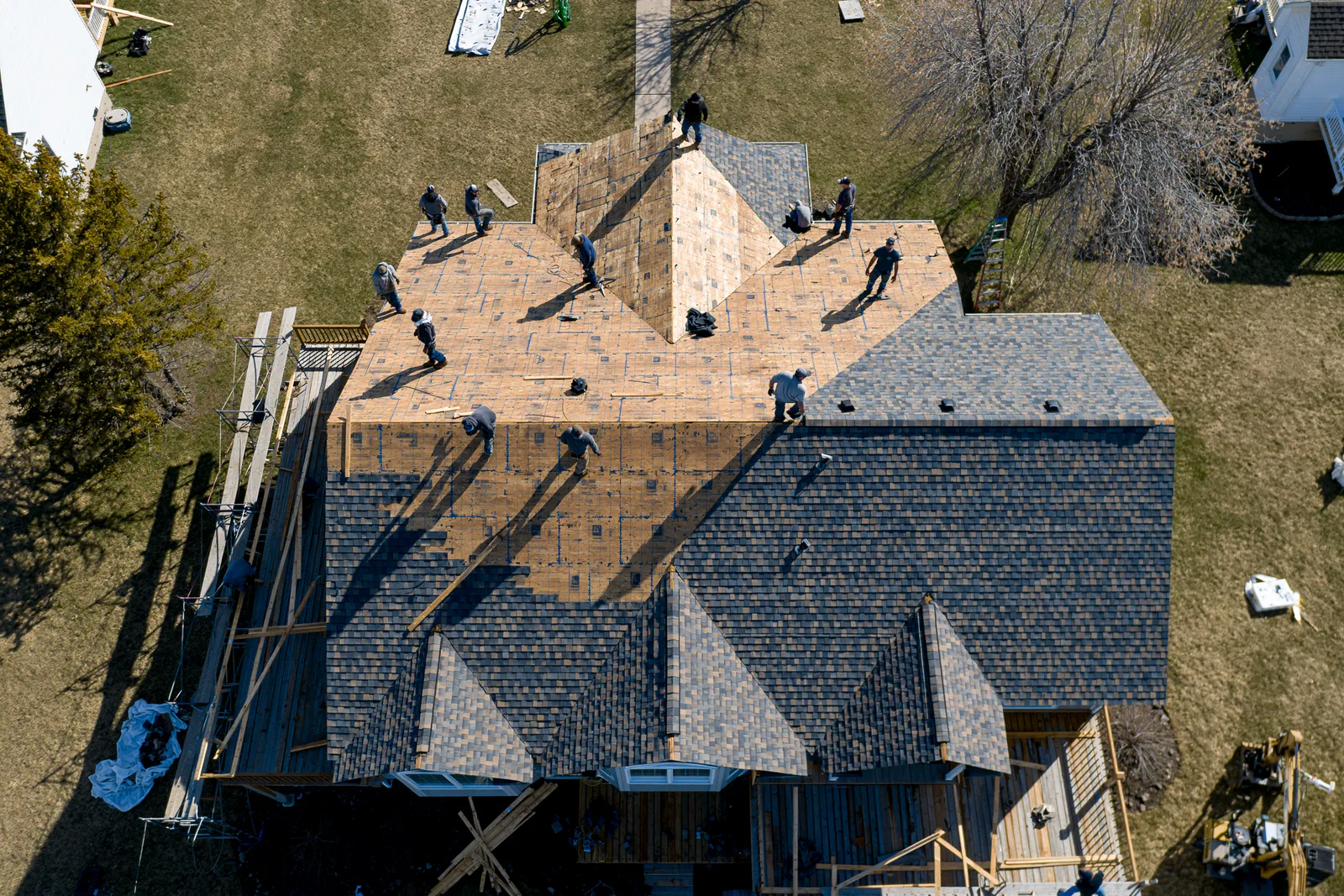 An aerial view shows roofers installing shingles across a large multi-gable roof