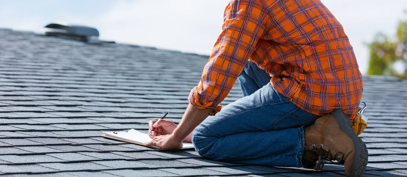 An inspector writing on a clipboard atop a gray shingle roof